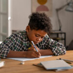 Criança debruçada em cadernos e livros em um ambiente doméstico, representando o ato de estudar em casa.