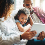 Uma criança sentada no sofá entre seus pais, observando a mãe apresentar o conteúdo de um tablet para ela. Como os pais influenciam a educação de seus filhos?