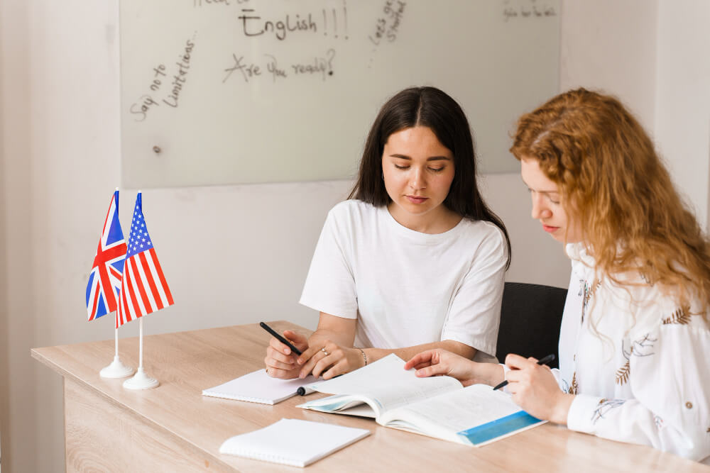 Duas adolescentes sentadas à mesa, olhando em direção a um livro aberto. Bandeiras dos Estados Unidos e da Grã Bretanha em cima da mesa, representando a prática de estudar inglês.