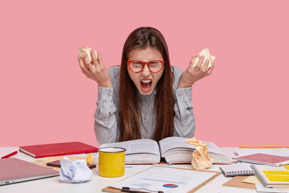 Jovem com livros abertos na sua frente, com papel amassado nas mãos e uma expressão de frustração, representando os desafios de lidar com a pressão no ensino médio.