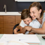 Mãe abraçando seu filho em frente a uma mesa com cadernos e folhas de papel, representando apoio para a tarefa de criar e seguir um plano de estudos.